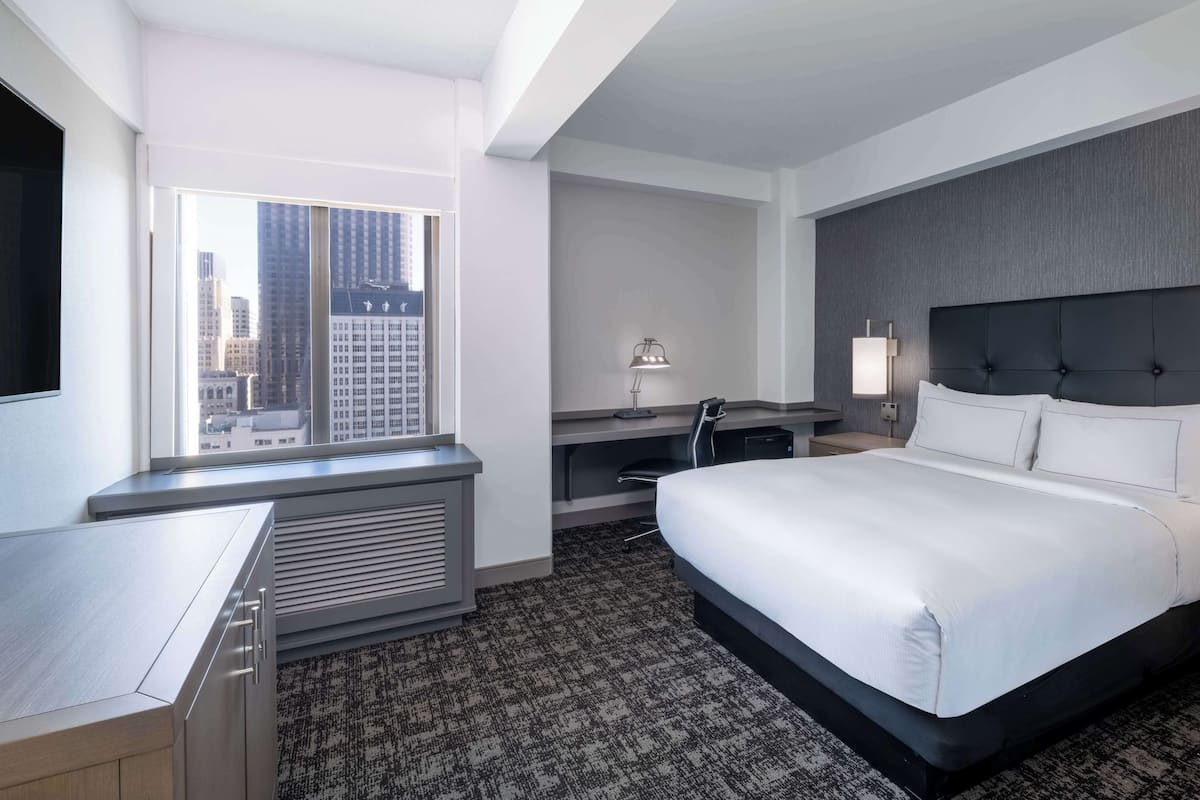 A hotel room with a large bed, a desk, a chair, a lamp, and a view of the cityscape through the window.