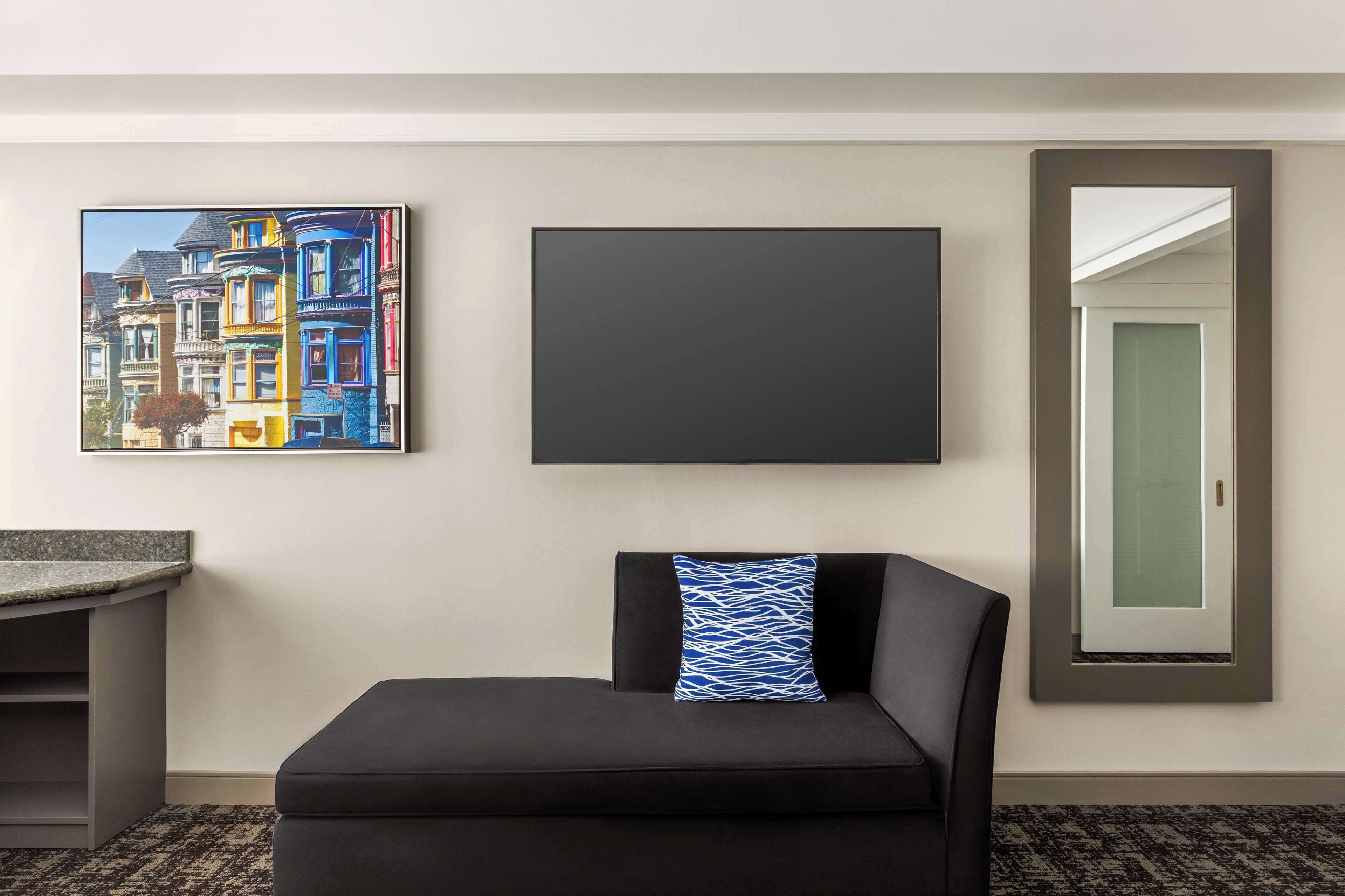 A hotel room with a black sofa, a framed picture of colorful buildings, a blank TV, and a mirror with a door.