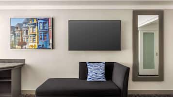 A hotel room with a black sofa, a framed picture of colorful buildings, a blank TV, and a mirror with a door.