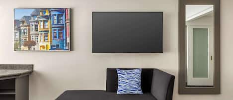 Habitación de hotel con sofá negro, un cuadro enmarcado de edificios coloridos, un televisor apagado y un espejo con puerta.