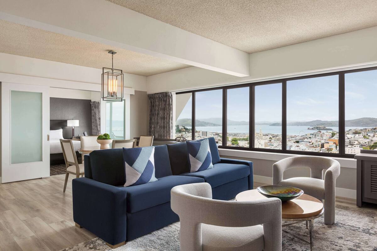 A modern living room with a blue sofa, a dining area, and a large window offering a city view.