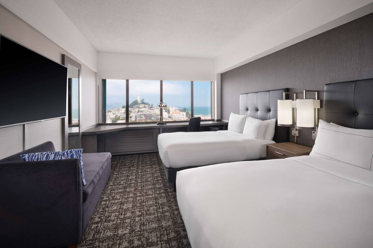 A hotel room with two beds, a large window with a city view, a flat-screen TV, and a desk with a chair.