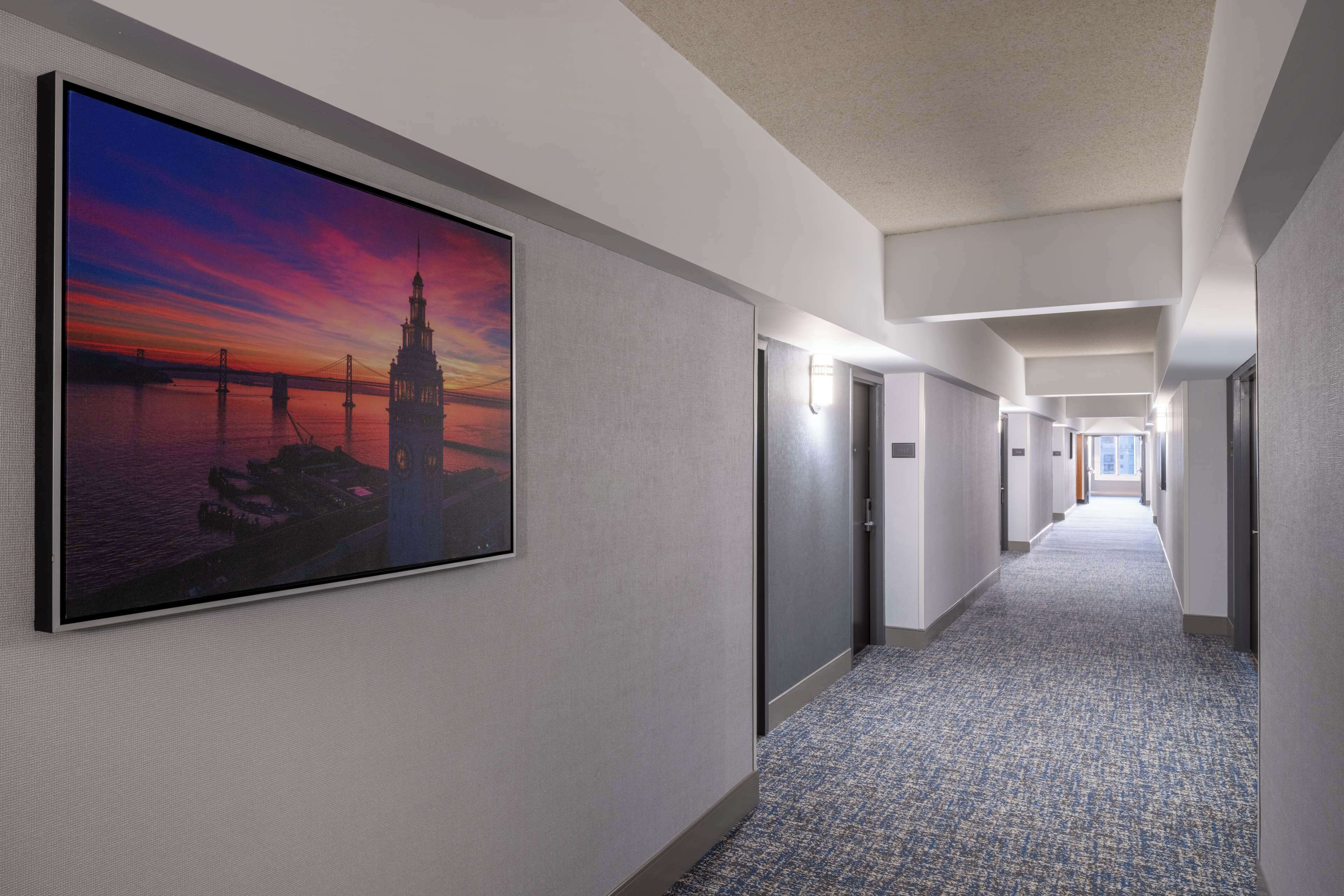A hotel hallway with a large framed picture of a sunset over a bridge and tower, carpeted floors, and doors leading to rooms.