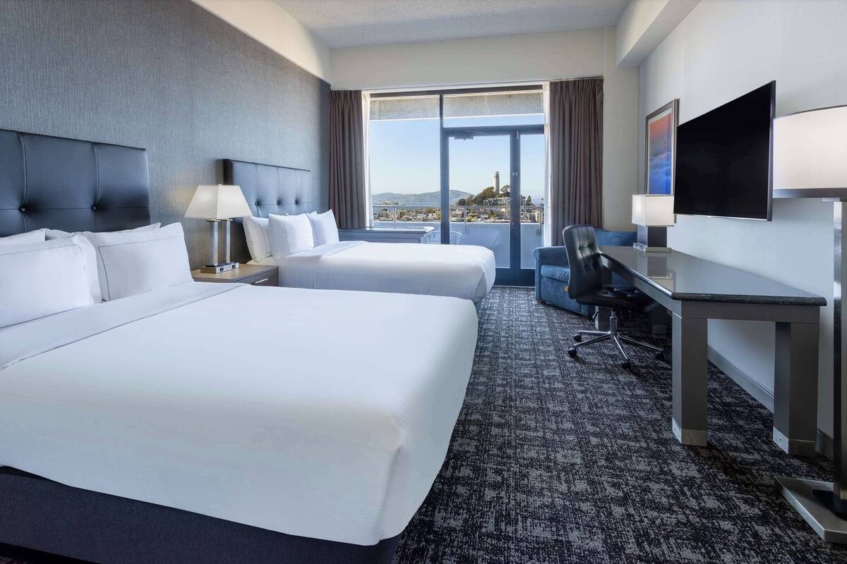A hotel room with two beds, a desk, a chair, a TV, and a large window offering a city view.