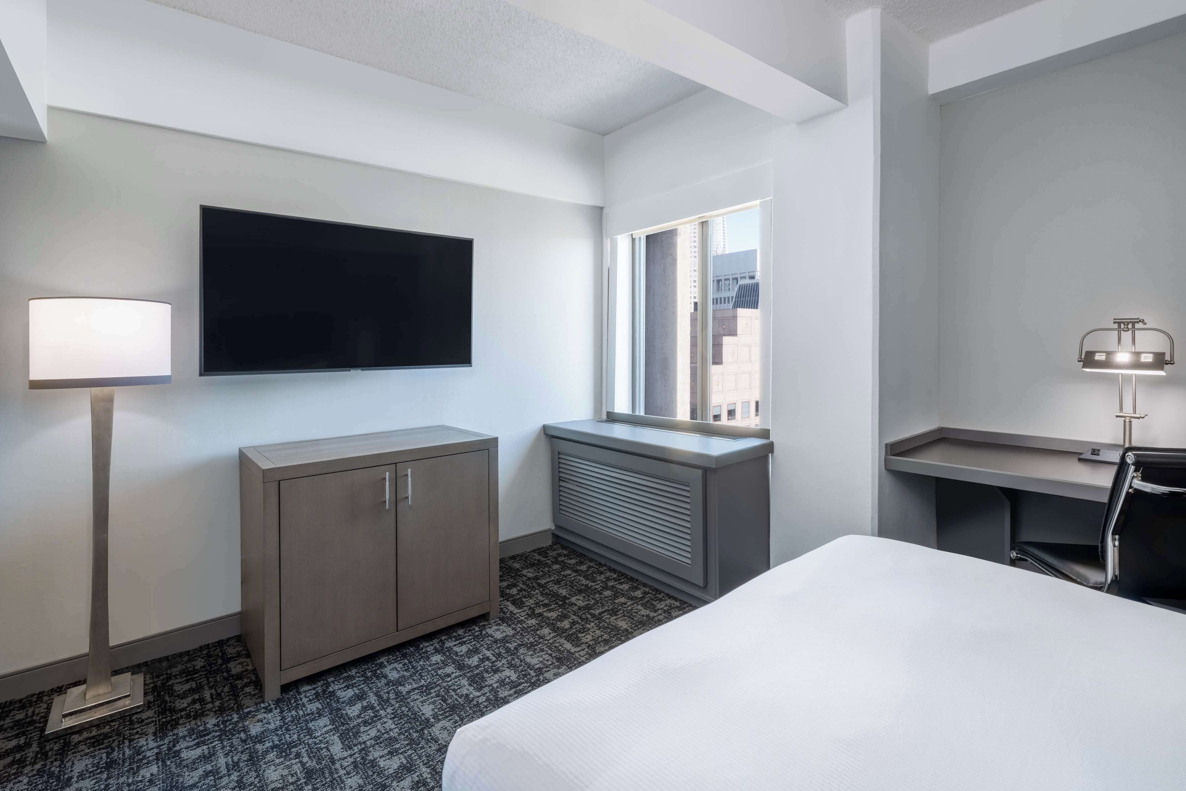 A hotel room with a bed, a TV mounted on the wall, a desk with a lamp, a wardrobe, and a window with a view of buildings.