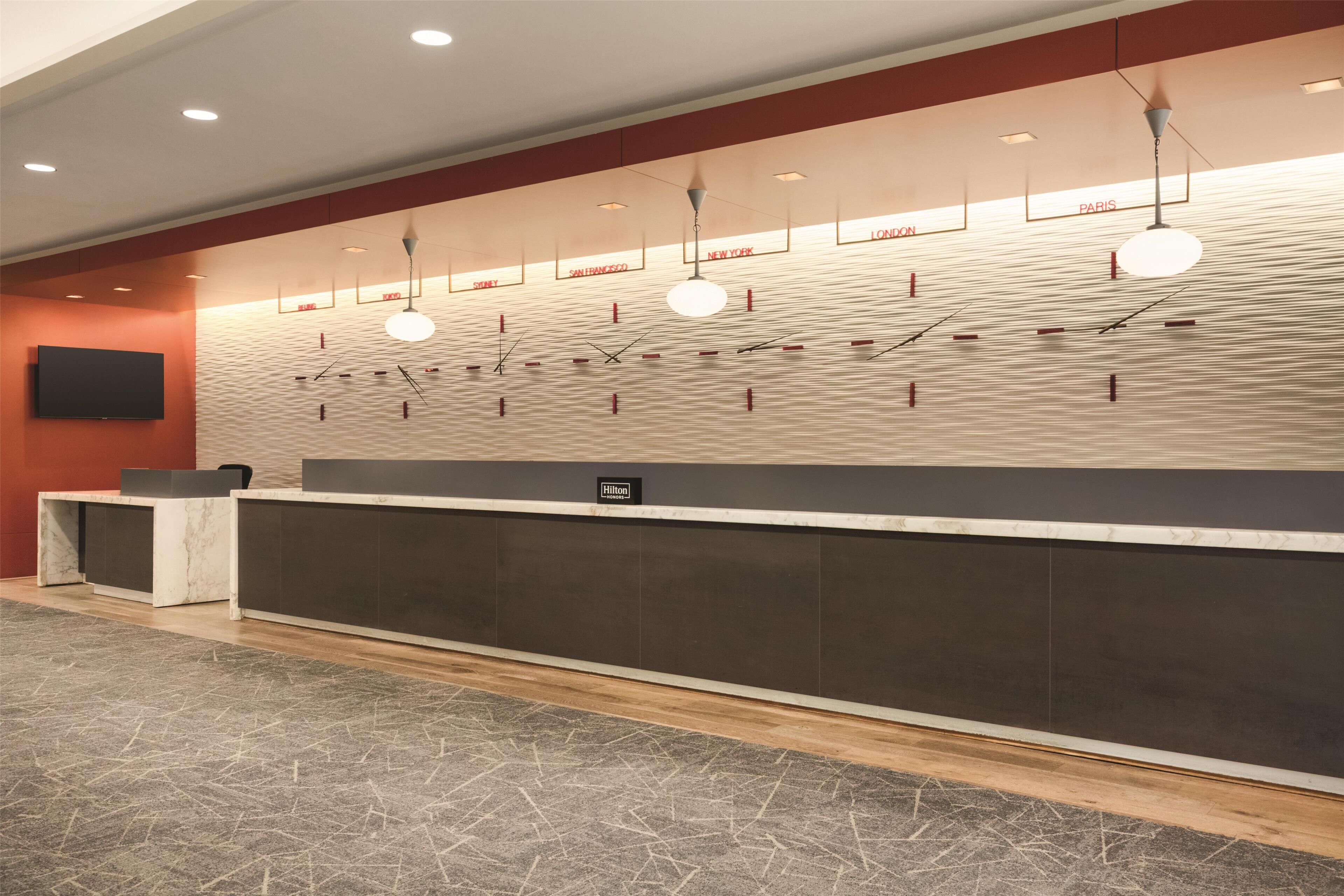 A hotel reception area with a long counter, a flat-screen TV, and a wall with city names.