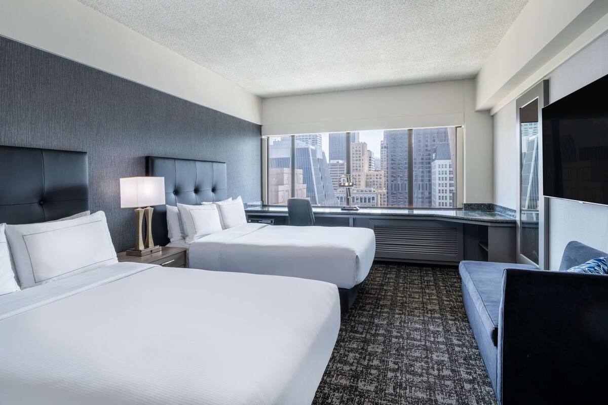 A hotel room with two beds, a large window with a city view, a desk, and a TV.