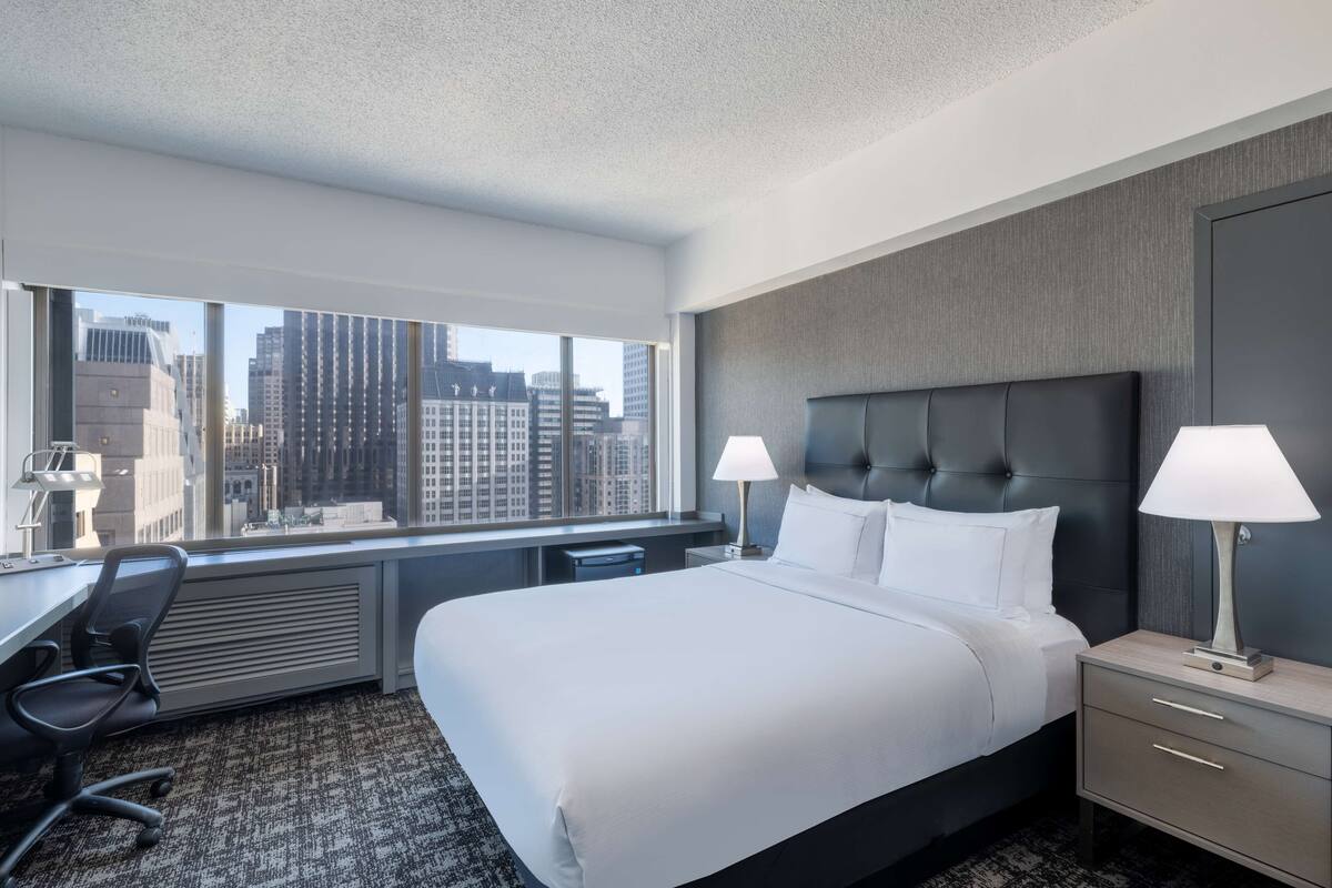 A hotel room with a large bed, two bedside lamps, a desk with a chair, and a view of the city through large windows.