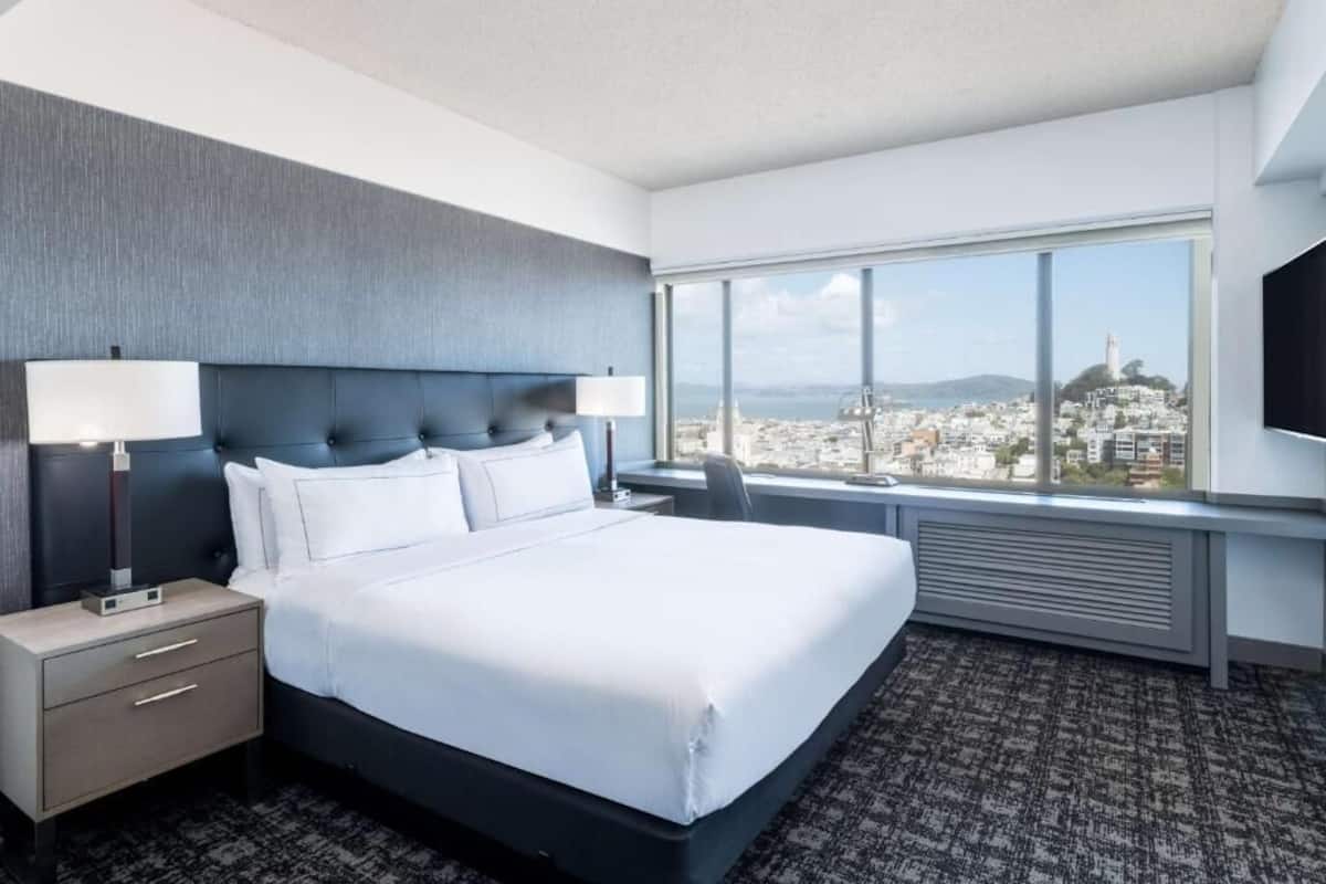 A hotel room with a large bed, a desk, a chair, a TV, and a window with a city view.
