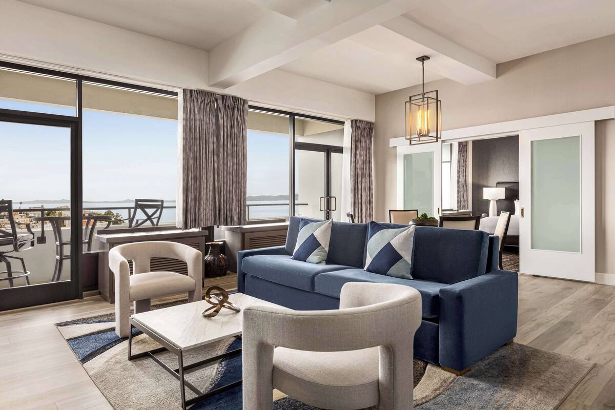 A modern living room with a blue sofa, two armchairs, a coffee table, and a view of the city through large windows.
