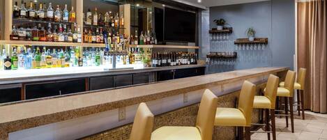 Una zona de bar con una barra alargada, varias estanterías llenas de botellas y varios taburetes de bar.