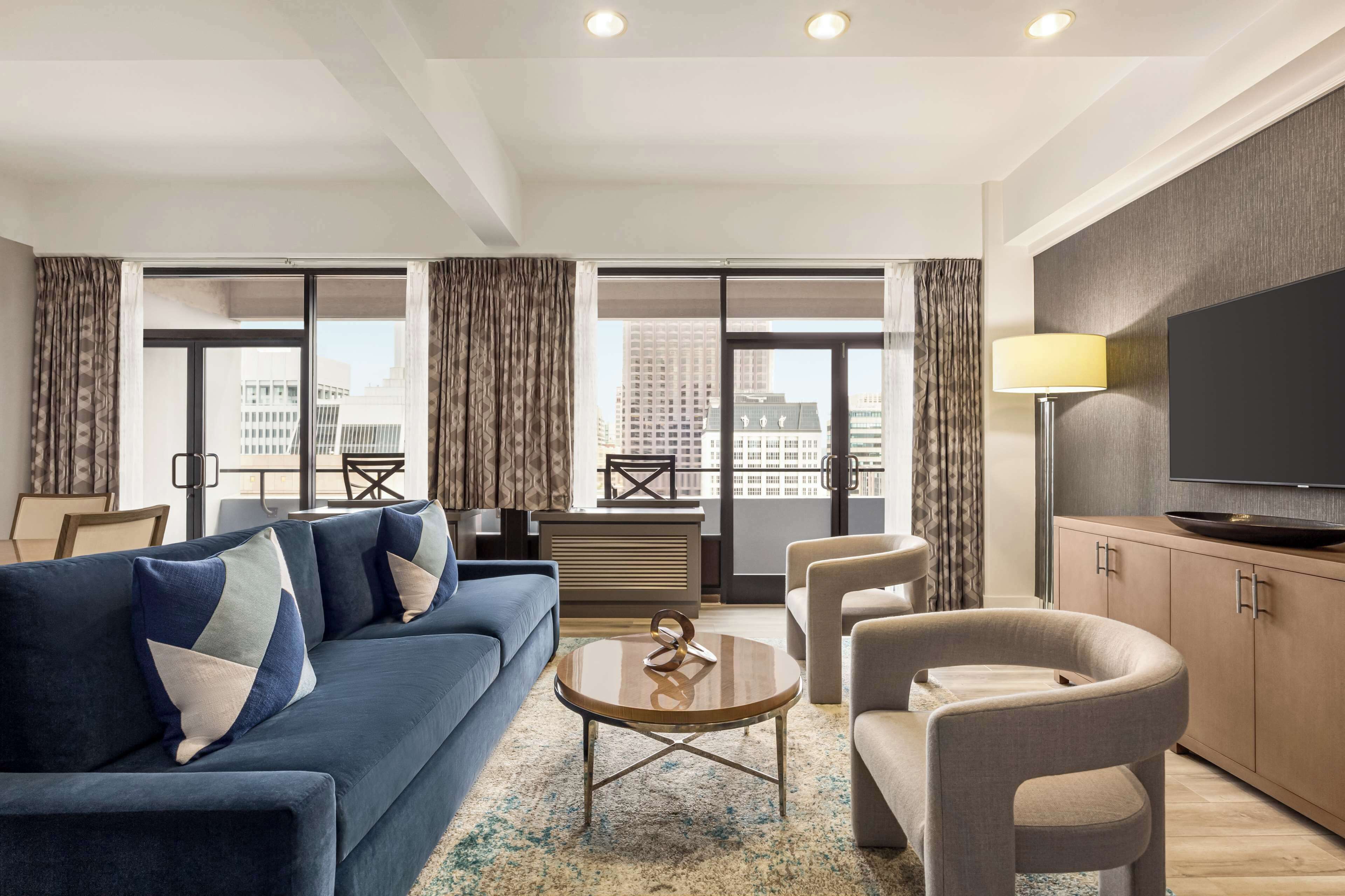 A modern hotel room with a blue sofa, a round coffee table, and a TV on a wooden cabinet.