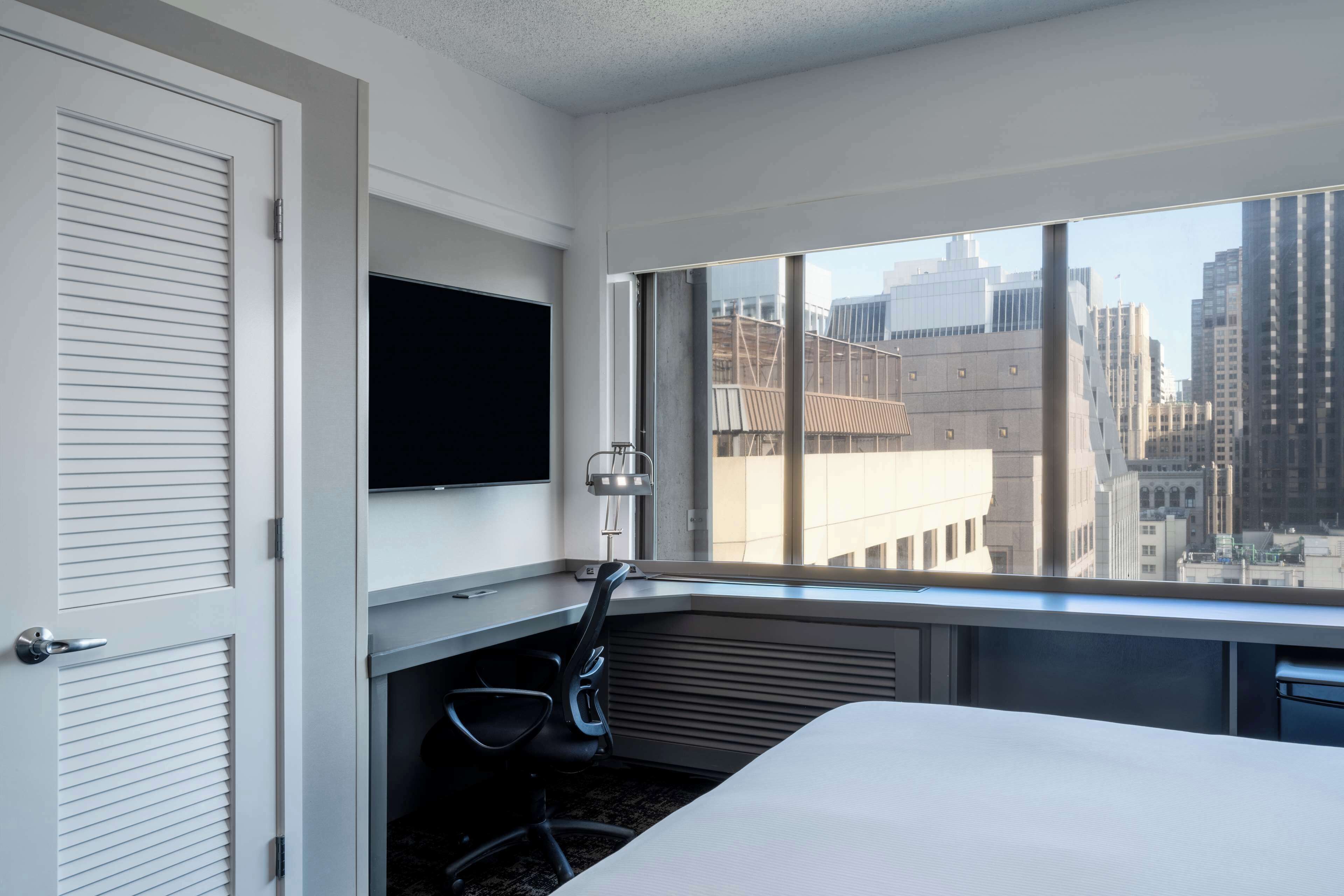 A hotel room with a large window, a flat-screen TV, a desk, and a chair.