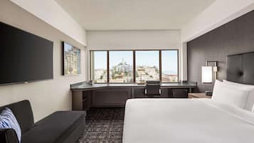 A hotel room with a large bed, a desk, a chair, a TV, and a window with a city view.
