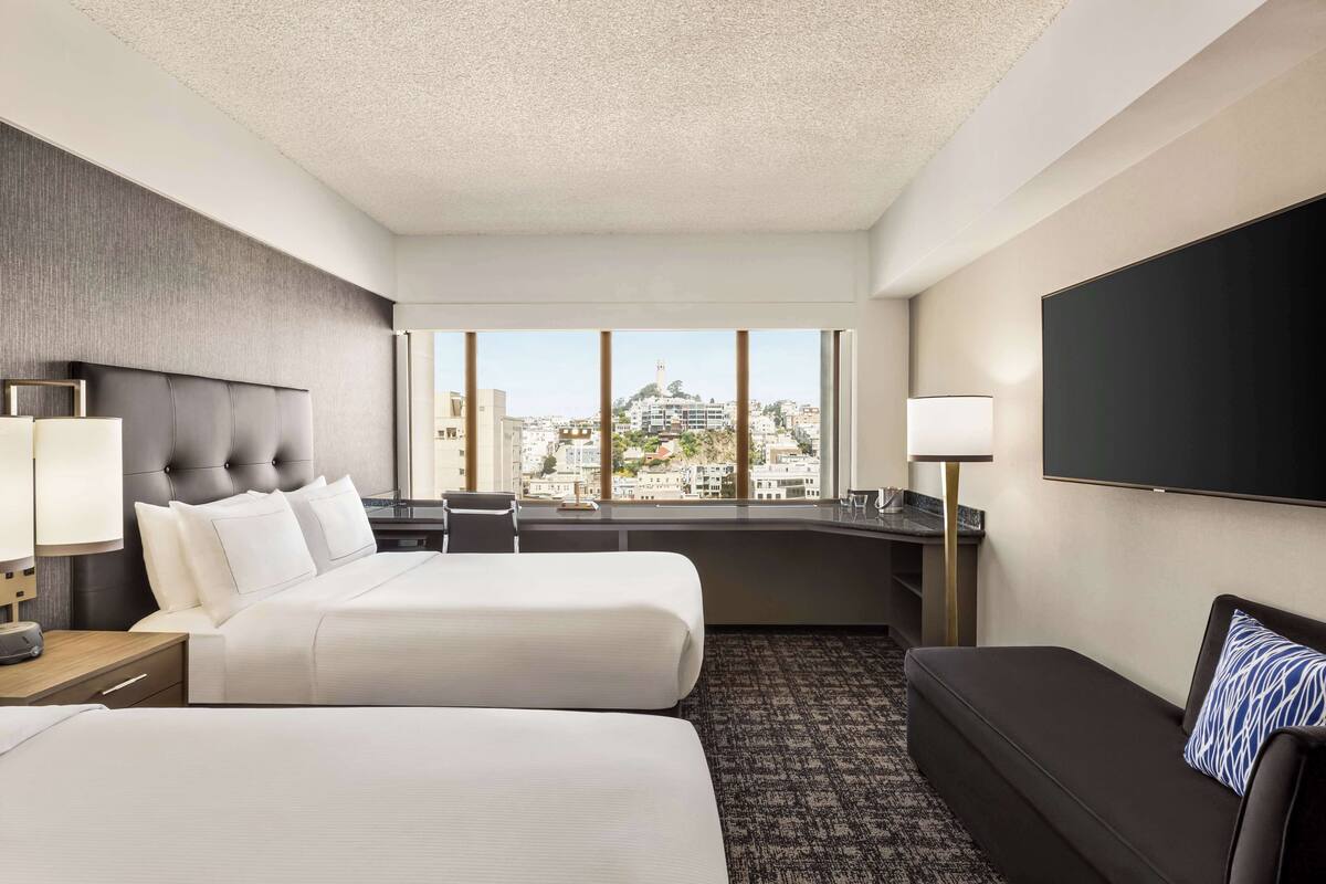 A hotel room with two beds, a sofa, a flat-screen TV, a large window with a city view, and a desk with a lamp.