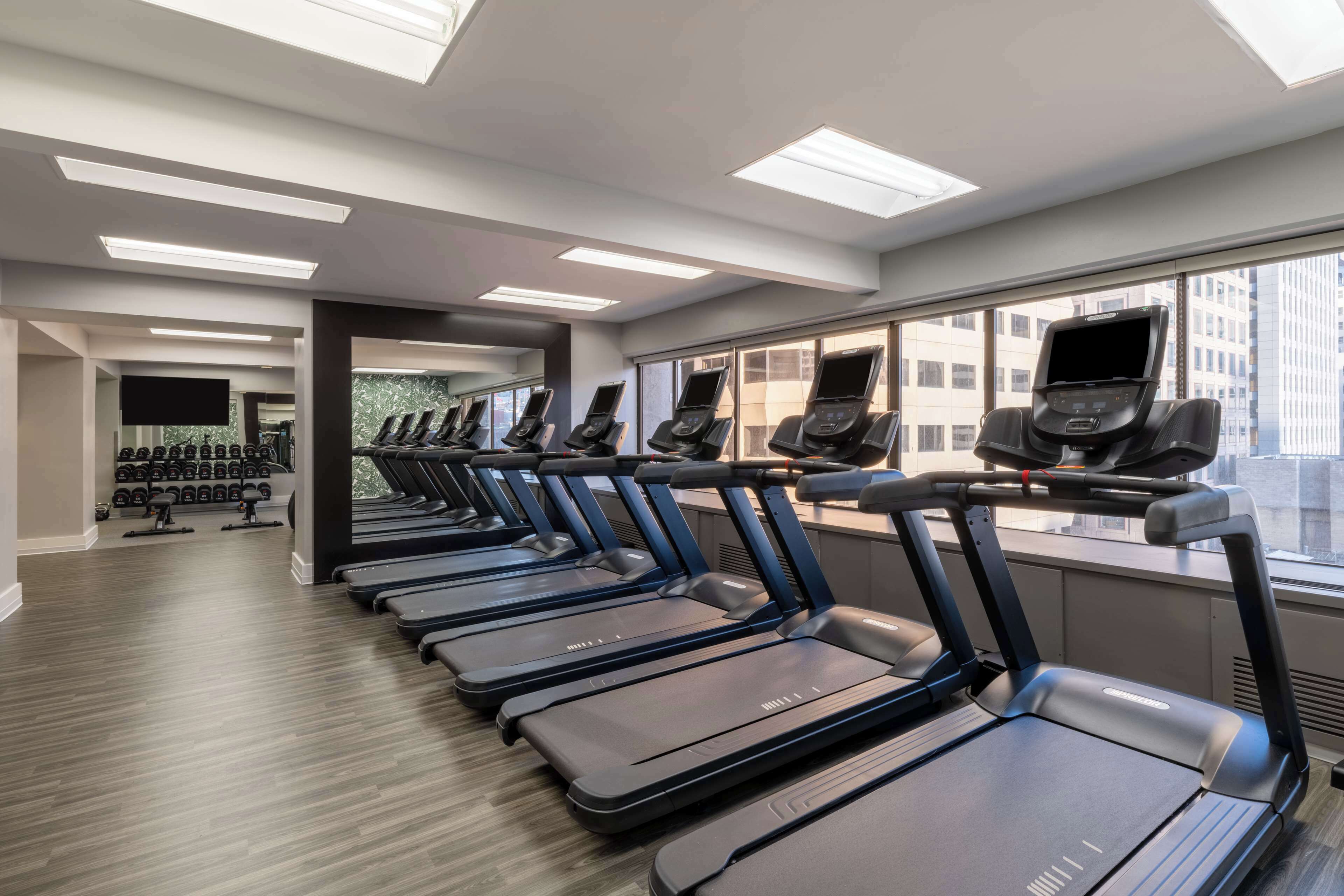 A row of treadmills in a gym with large windows and a view of buildings.