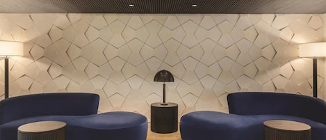 Un área de descanso moderna con una pared de diseño geométrico, dos sofás azules y dos mesitas redondas.