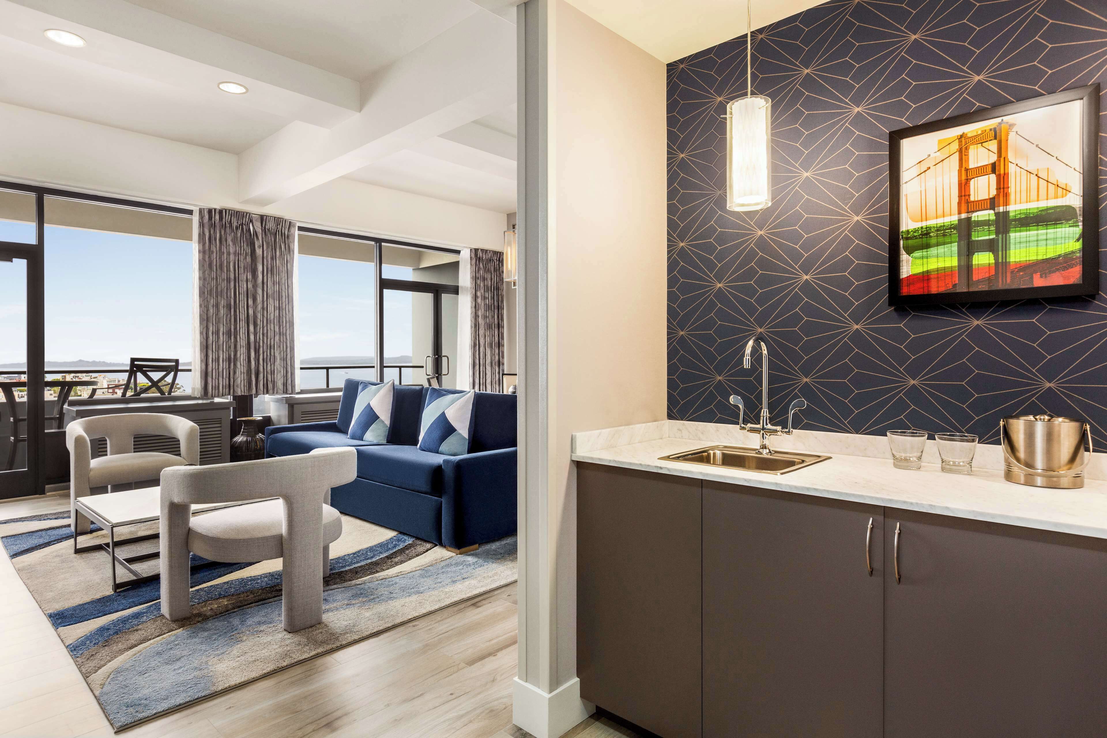 A modern hotel room with a large window, a sofa, a dining area, and a kitchen with a sink and countertop.