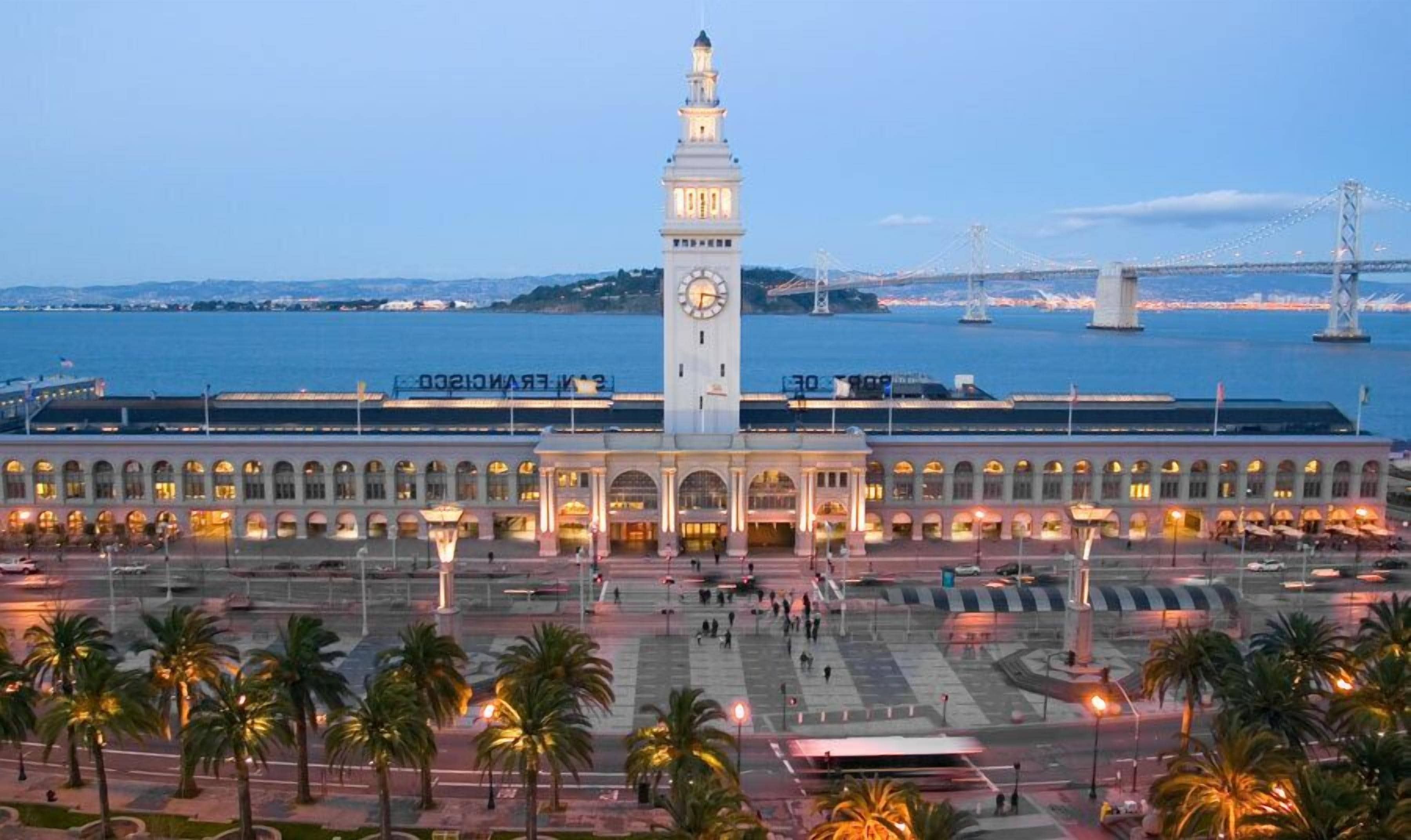 A large, historic building with a prominent clock tower, a spacious plaza with palm trees, and a waterfront view.