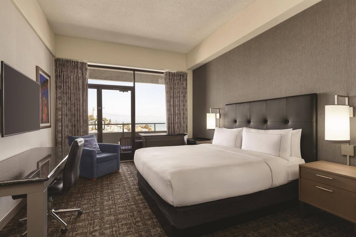 A hotel room with a large bed, a desk, a chair, a TV, and a window with curtains.