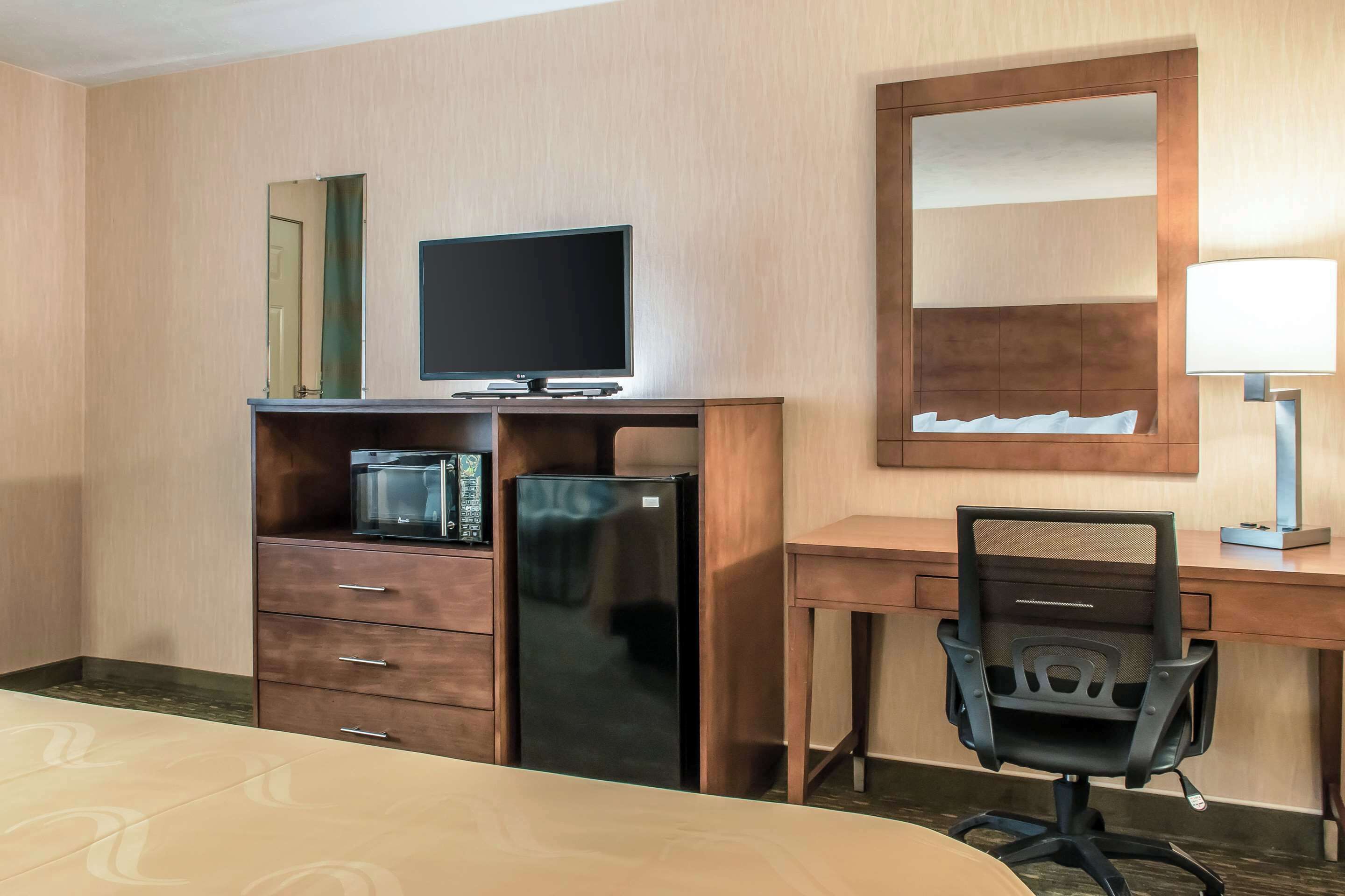 A hotel room with a bed, a desk with a computer, a refrigerator, a microwave, a chair, and a large mirror.