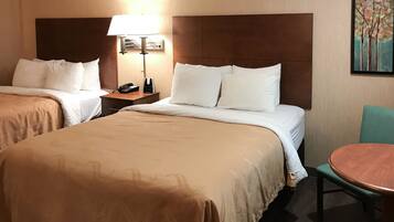 A hotel room with two beds, a bedside table with a lamp, a small table with a chair, and a painting on the wall.