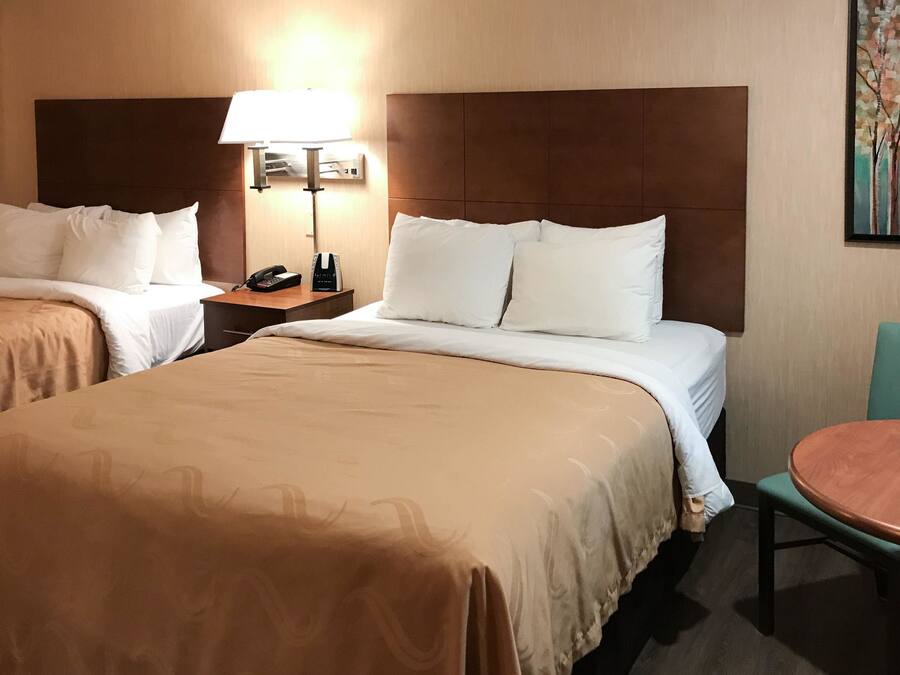 A hotel room with two beds, a bedside table with a lamp, a small table with a chair, and a painting on the wall.