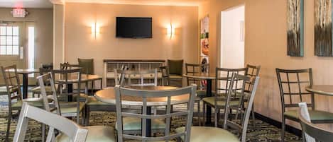 A hotel room with a large round table, multiple chairs, a flat-screen TV mounted on the wall, and a patterned carpet.