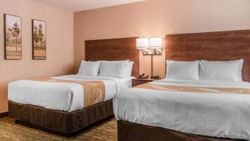 A hotel room with two beds, a wooden headboard, a nightstand with a lamp, and two framed pictures on the wall.