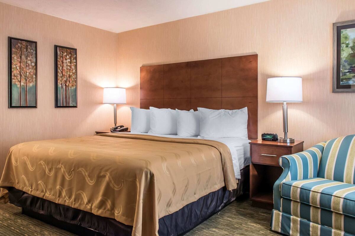 A hotel room with a large bed, two bedside lamps, a nightstand, a striped armchair, and two framed paintings on the wall.