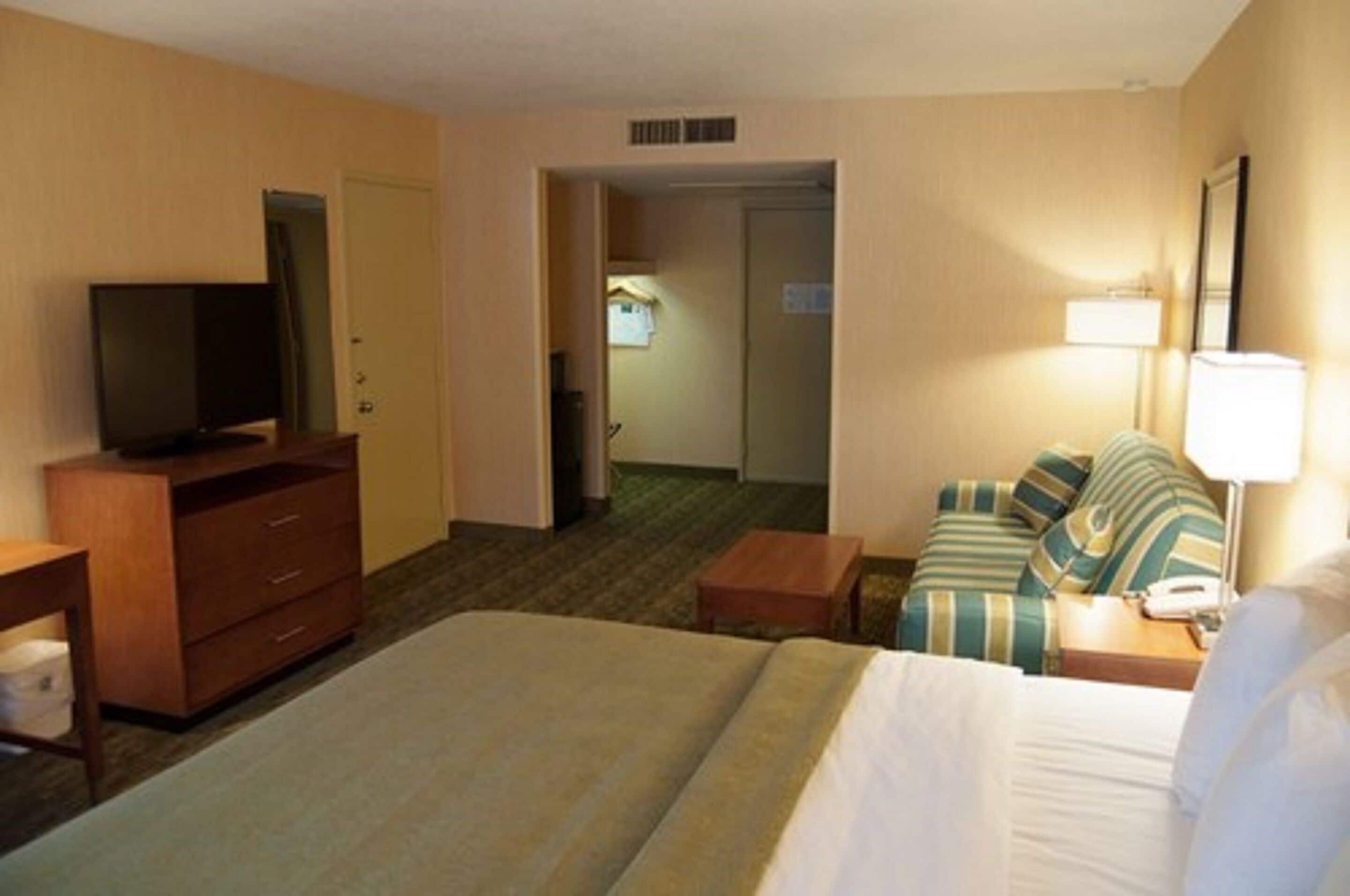 Habitación de hotel con cama, televisión sobre una cómoda de madera, sofá con almohadas a rayas y mesita de noche con lámpara.
