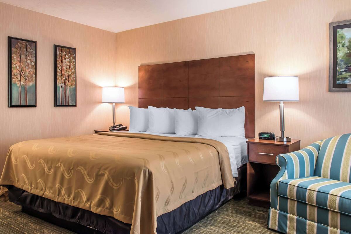 A hotel room with a large bed, two bedside lamps, a nightstand, a striped armchair, and two framed paintings on the wall.