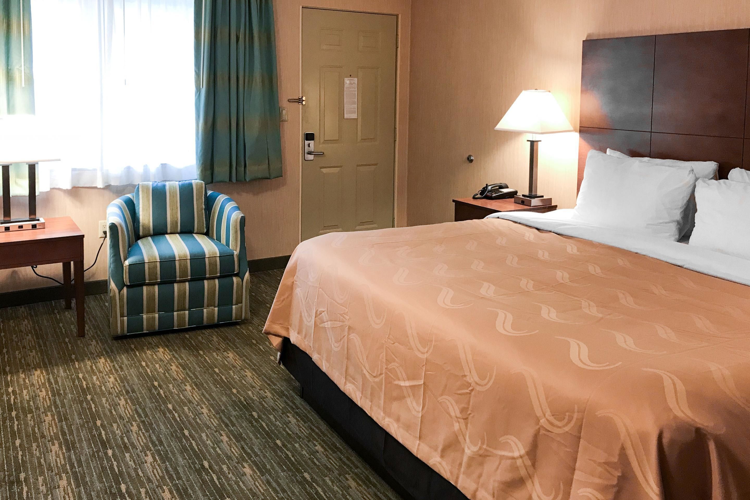 A hotel room with a bed, a striped armchair, a desk, a lamp, and a door.