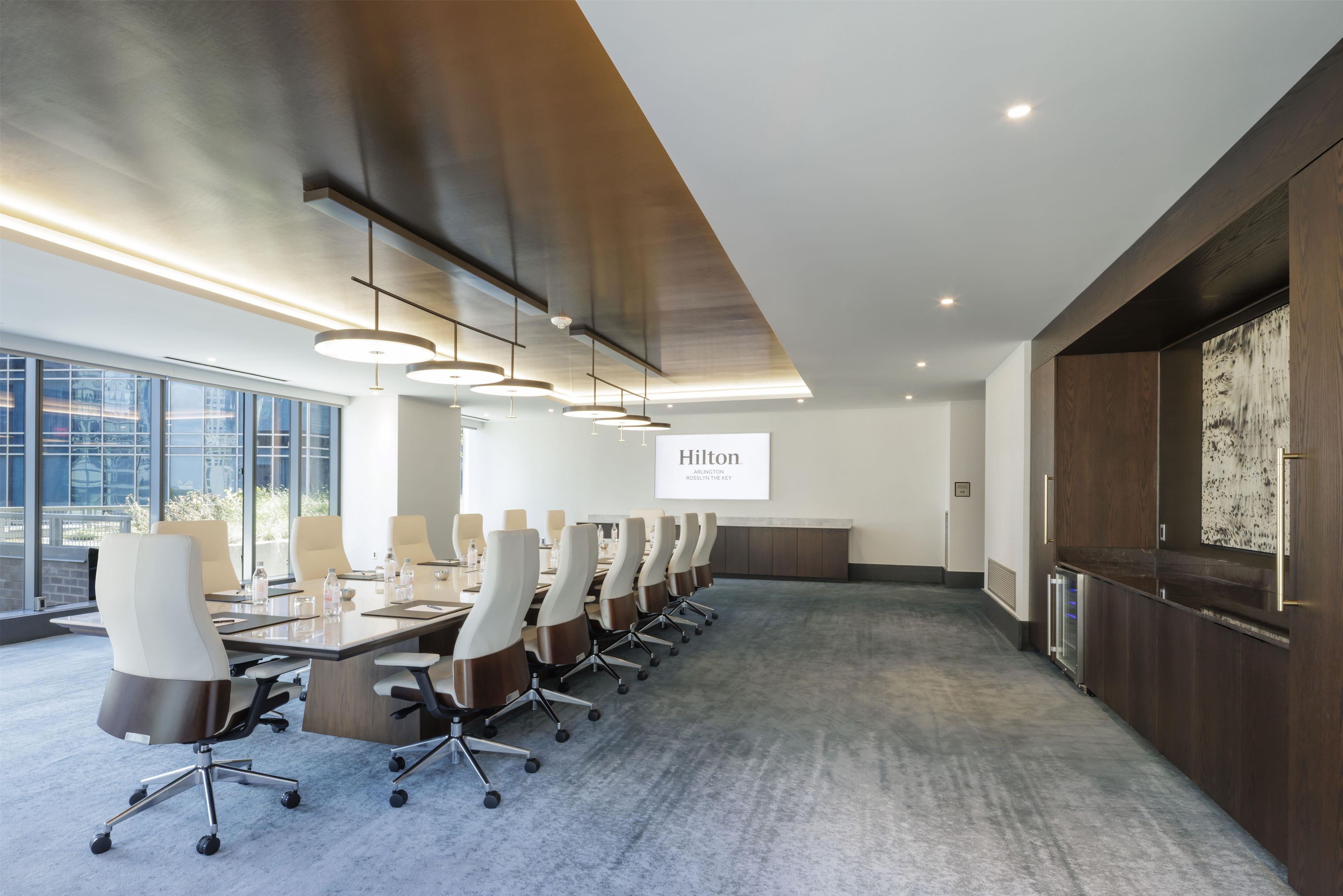 A modern conference room with a long table, chairs, and a projector screen displaying 'Hilton Arlington Rosslyn The Key'.