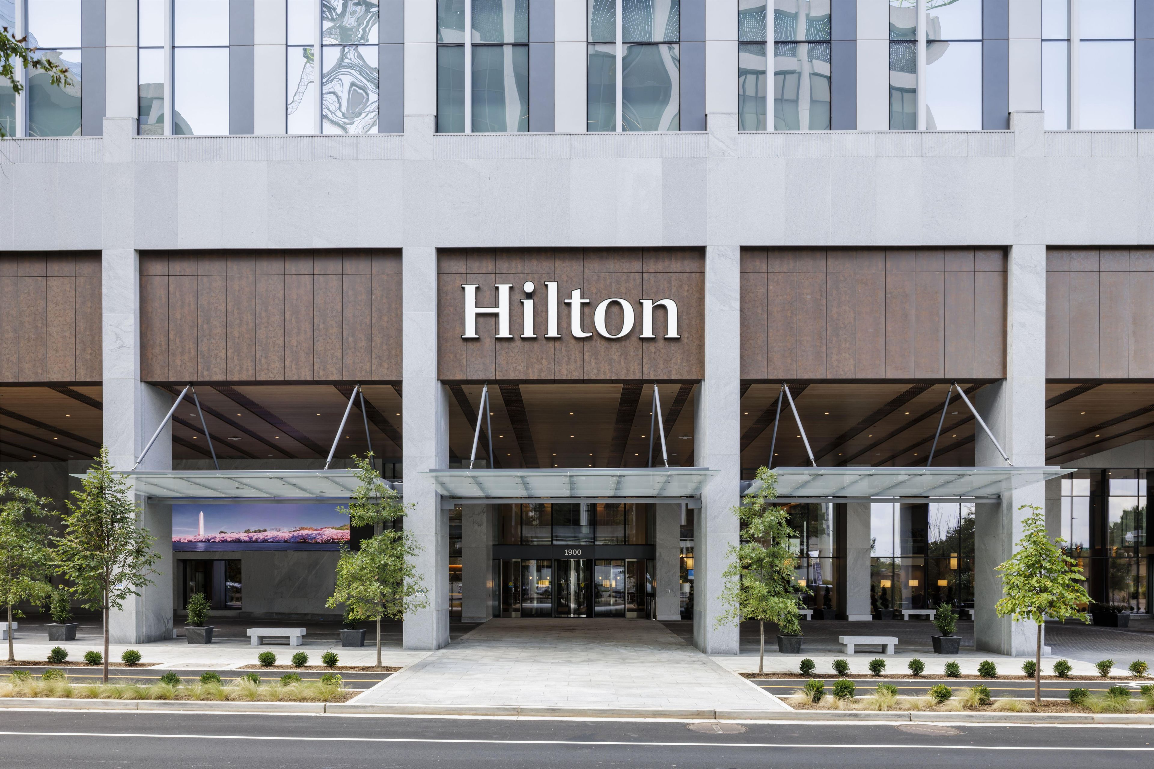 A modern hotel entrance with a prominent 'Hilton' sign, covered entryway, and landscaped surroundings.