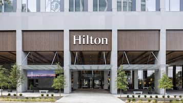 A modern hotel entrance with a prominent 'Hilton' sign, covered entryway, and landscaped surroundings.