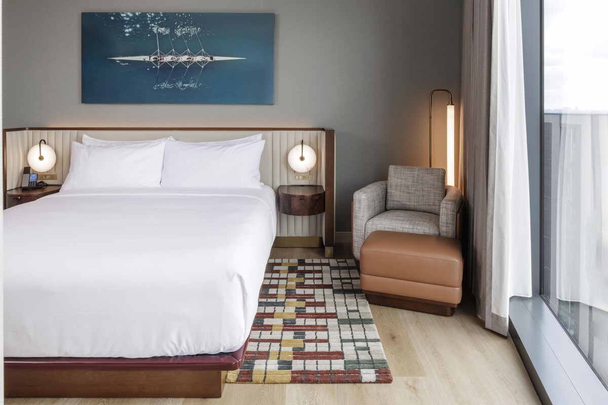 A hotel room with a large bed, a chair, a nightstand, a lamp, and a window with curtains.