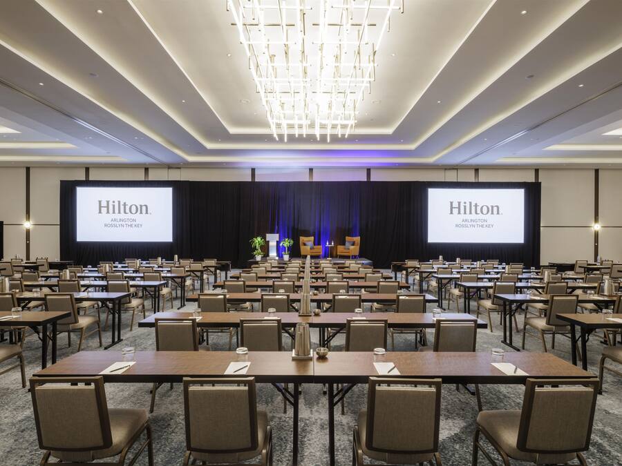 A large conference room with rows of tables and chairs, a stage with a large screen displaying the Hilton logo, and a modern chandelier.