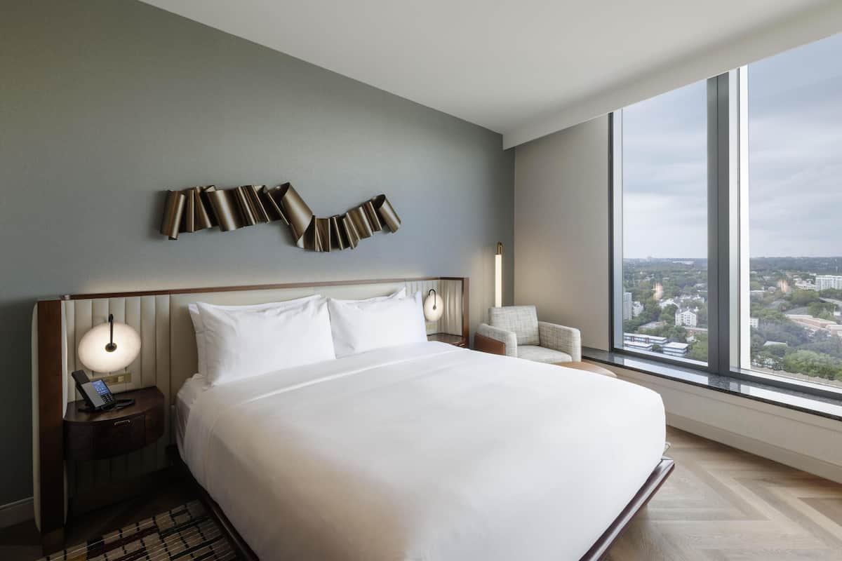 Une chambre d’hôtel moderne avec un grand lit, une table de chevet avec une lampe, un fauteuil et une vue sur la ville grâce à de grandes fenêtres.