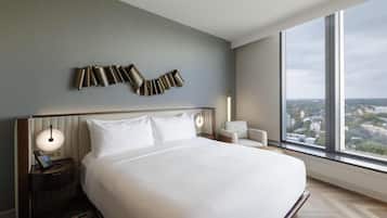 A hotel room with a large bed, a nightstand with a phone, a lamp, and a view of the city through large windows.