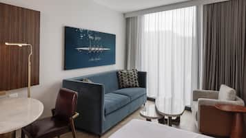 A modern hotel room with a blue sofa, a round dining table, a small side table, and a large window with curtains.