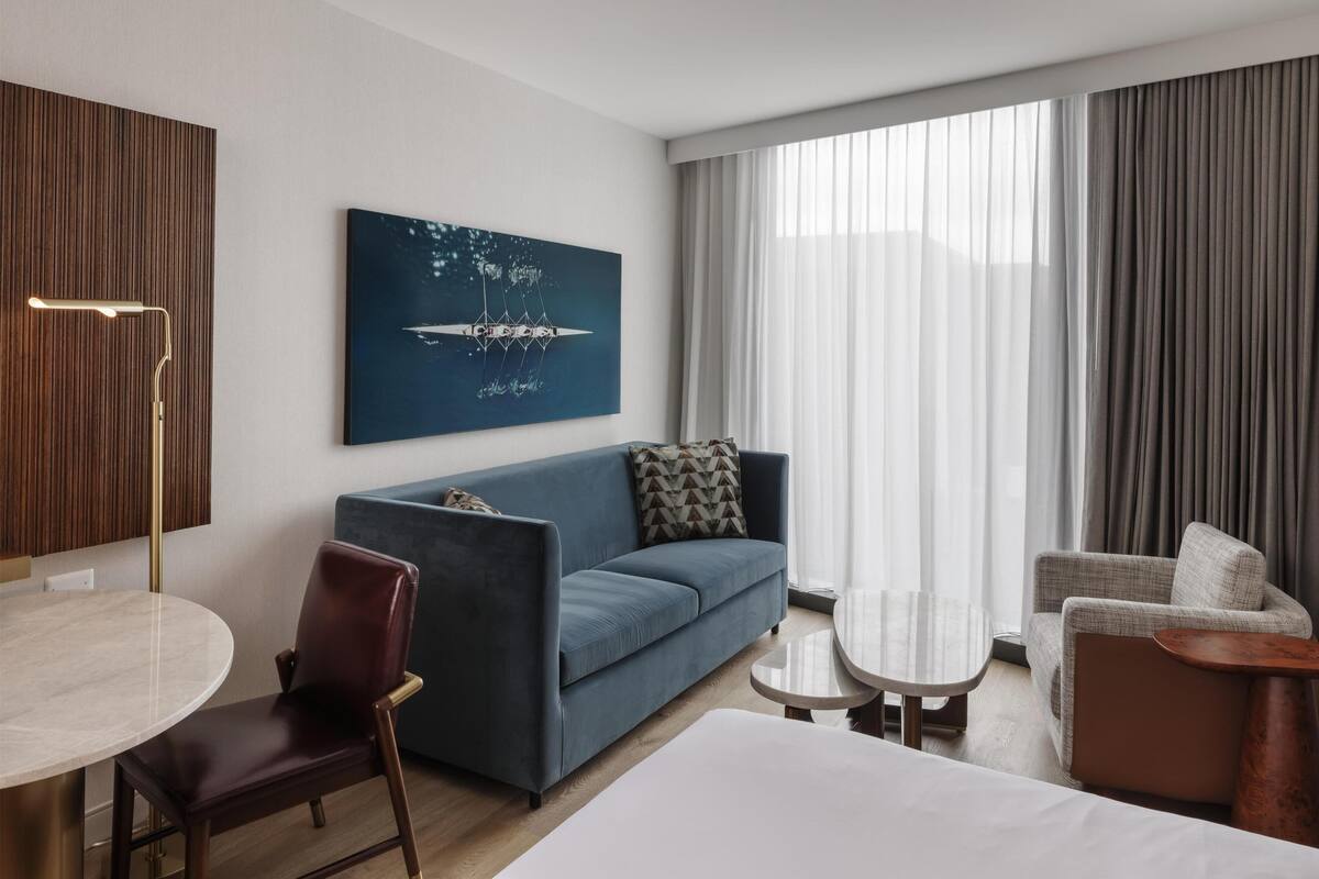 Une chambre d’hôtel moderne avec un canapé bleu, une table de salle à manger ronde, une petite table d’appoint et une grande fenêtre avec des rideaux.