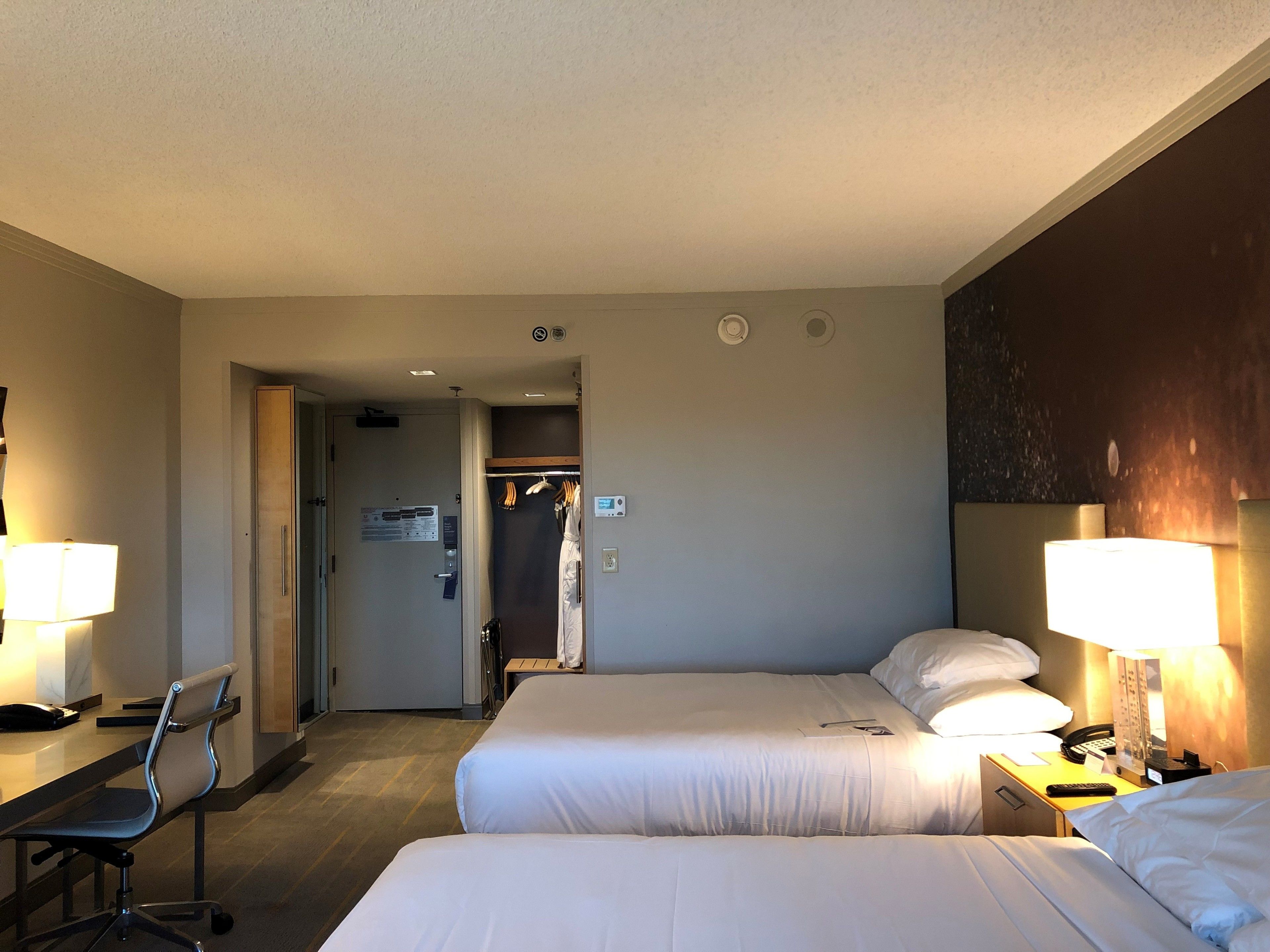 Ein Hotelzimmer mit zwei Betten, einem Schreibtisch mit Stuhl, einem Kleiderschrank und einem durch eine offene Tür einsehbaren Badezimmer.