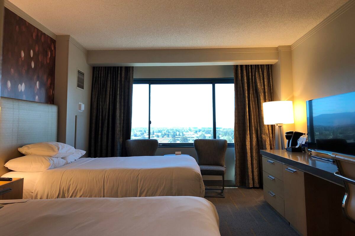 A hotel room with two beds, a desk with a flat-screen TV, a chair, and a large window offering a city view.