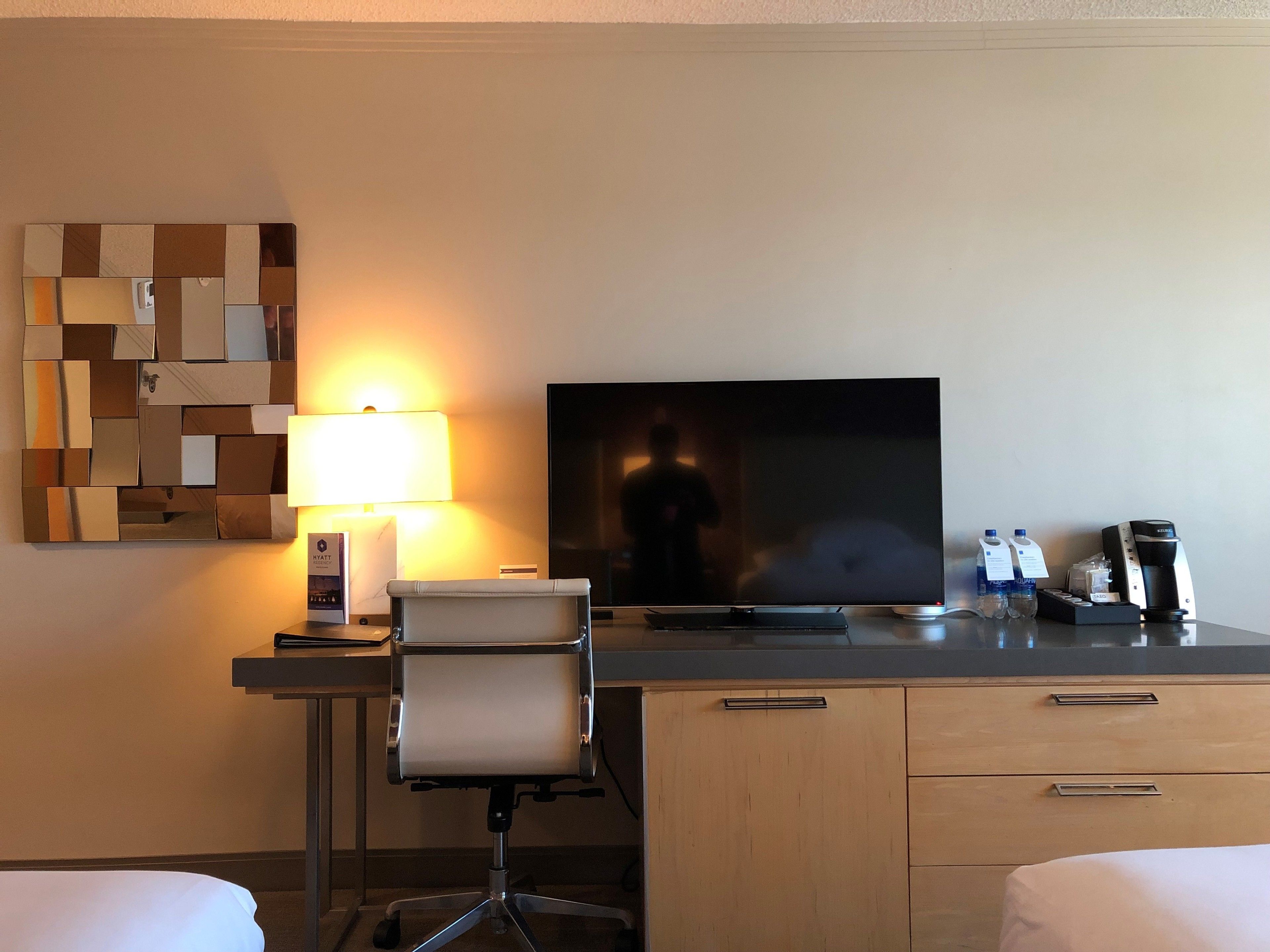 Ein Hotelzimmer mit Schreibtisch, Stuhl, Fernseher, Lampe, Kaffeemaschine und einer Minibar.