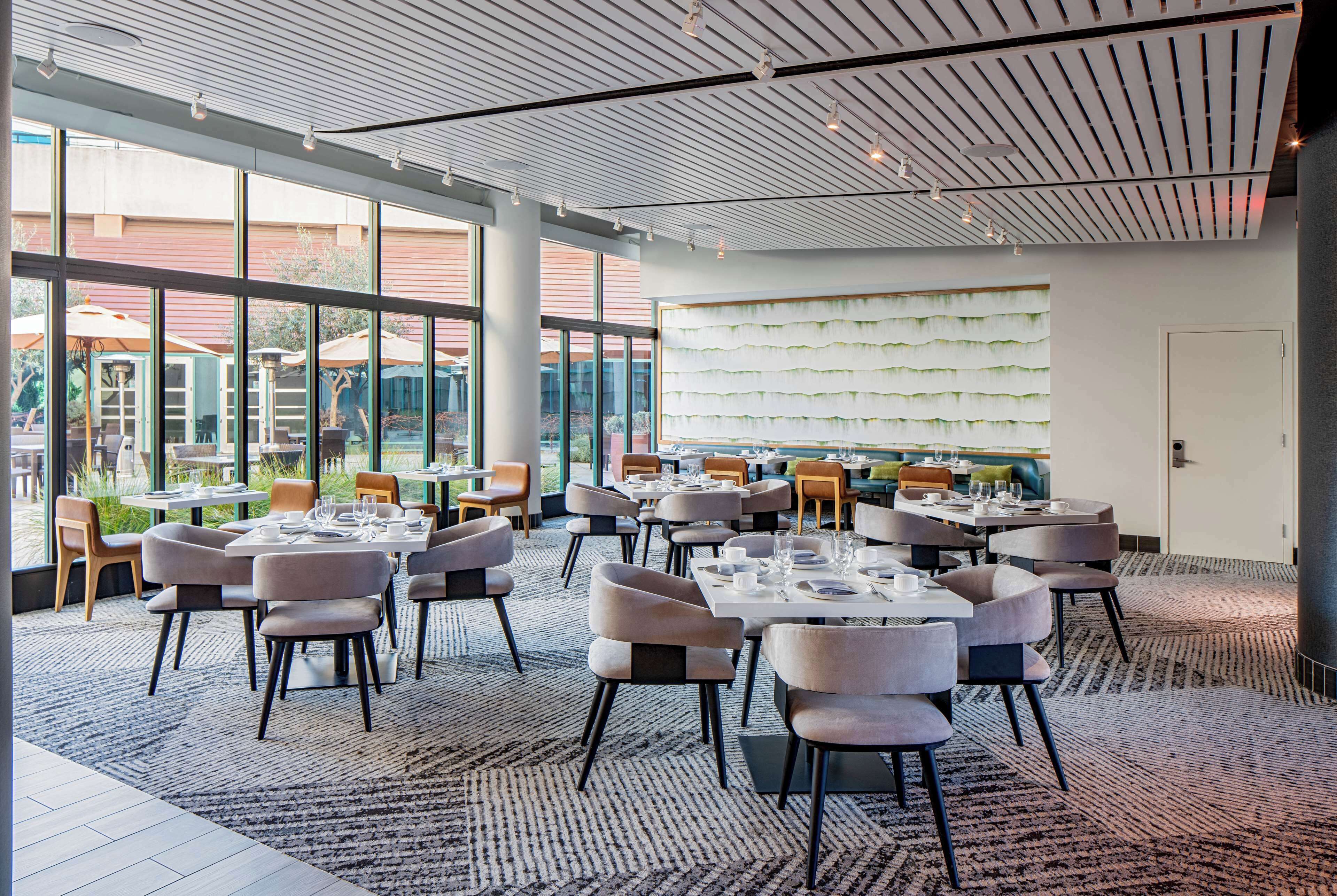A spacious dining area with multiple tables and chairs, large windows offering views of the outdoors, and a modern ceiling design.