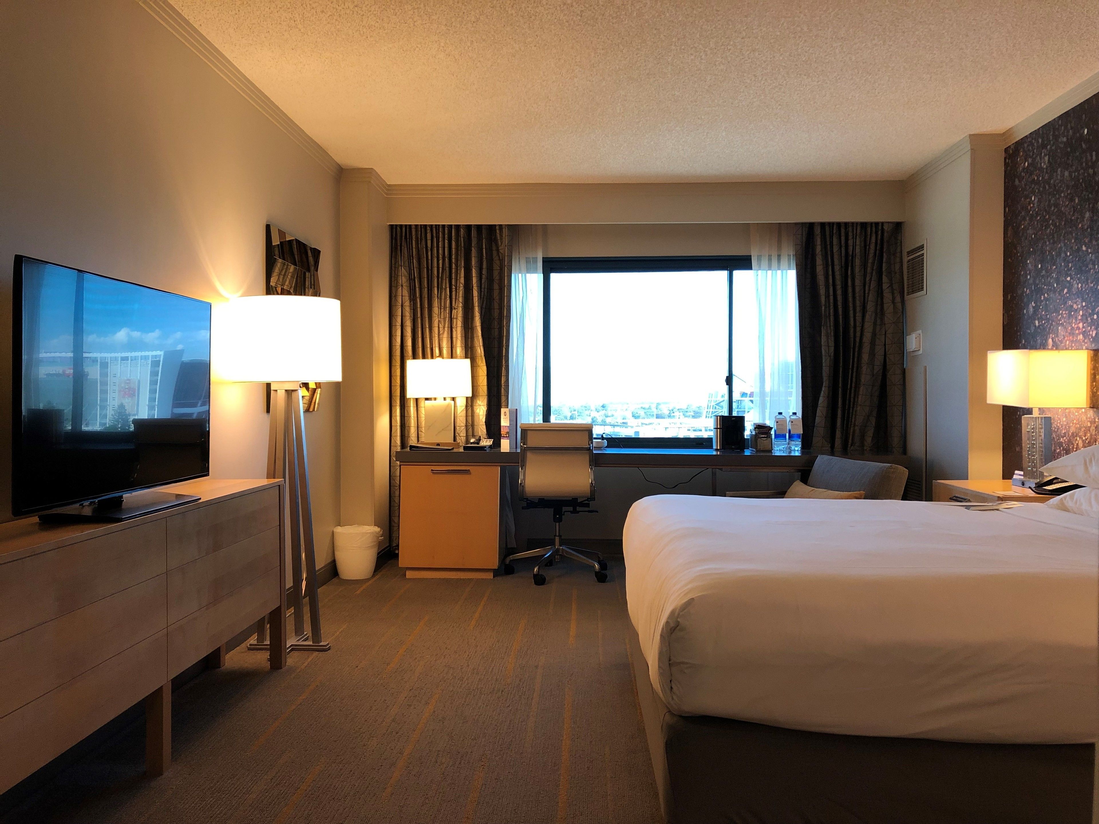 A hotel room with a bed, desk, chair, TV, and lamps.