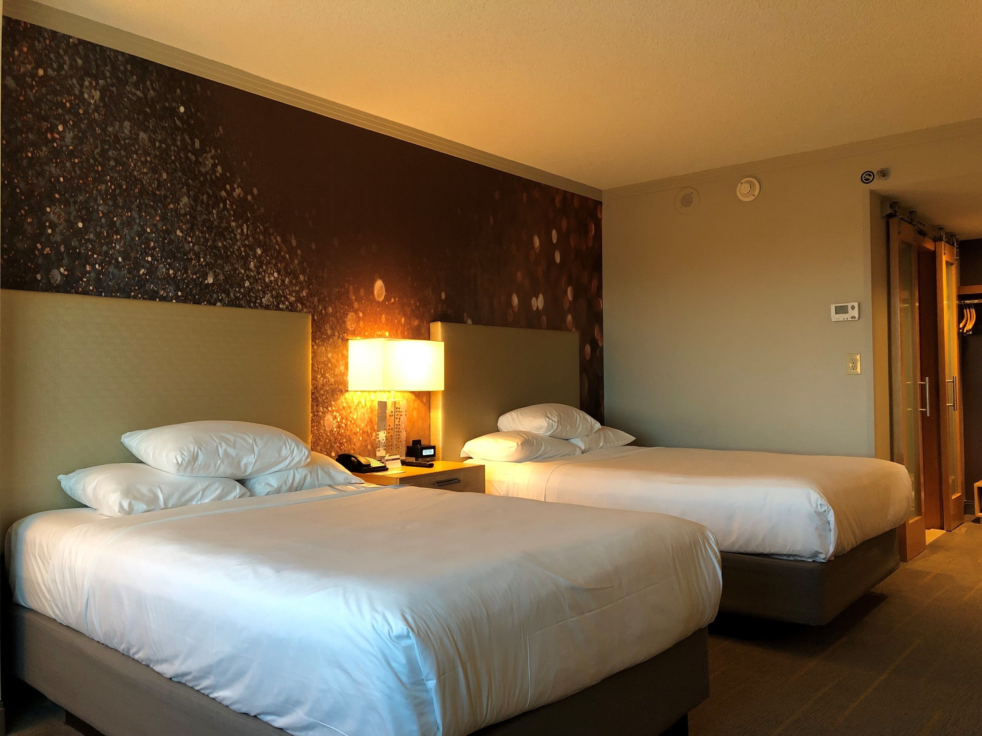 A hotel room with two beds, a nightstand with a lamp, a phone, and a thermostat on the wall.