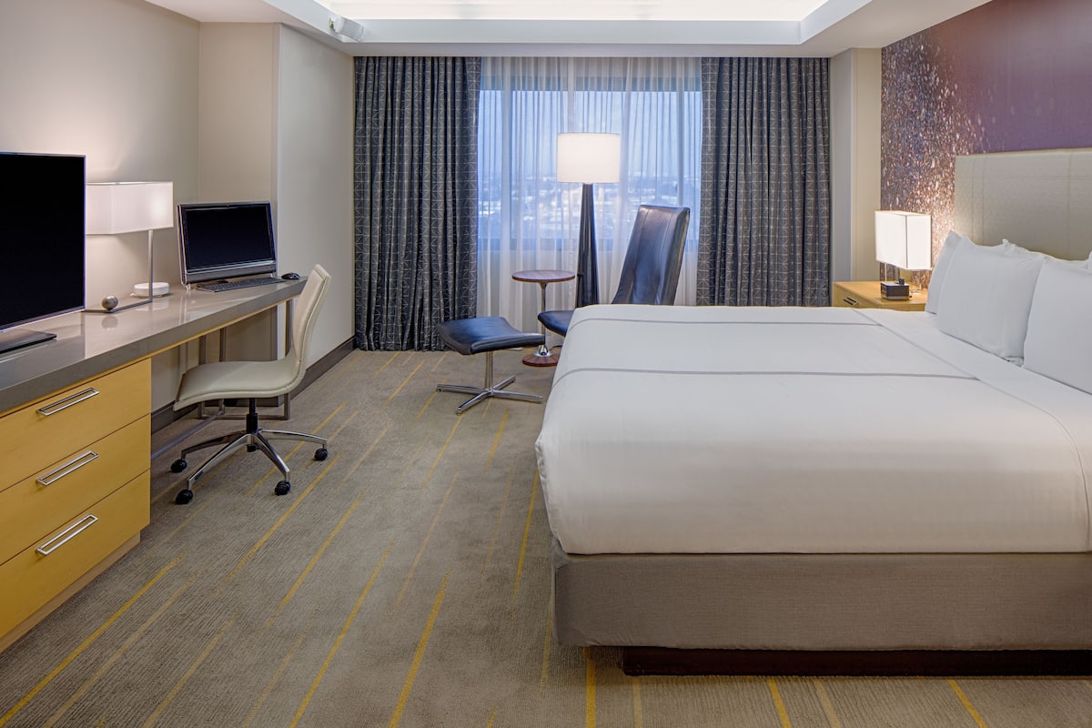 A hotel room with a large bed, a desk with a computer, a chair, a small table, and a TV.