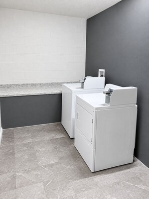 A laundry room with two white washing machines, a grey tiled floor, and a white tiled wall. - Super 8 by Wyndham Hillsville (Hillsville)